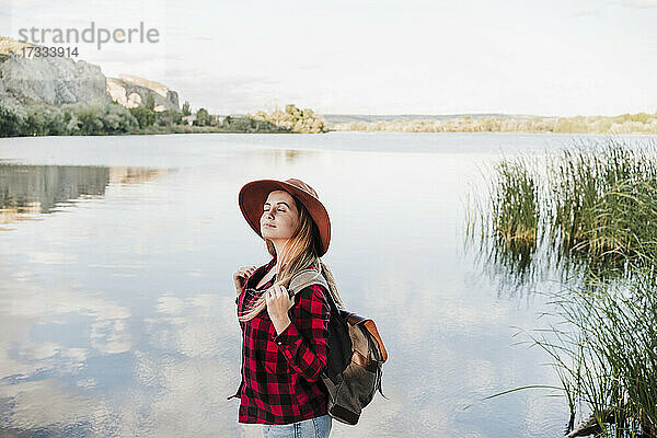 Junge schöne Frau mit Hut steht mit geschlossenen Augen am Seeufer