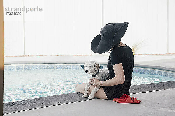 Frau mit Hut streichelt Hund  während sie im Urlaub am Pool sitzt