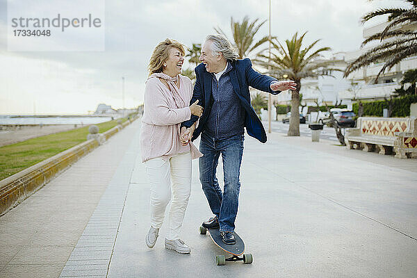 Fröhlicher reifer Mann  der mit dem Skateboard an einer Frau vorbeifährt  die sich auf dem Gehweg an den Händen hält