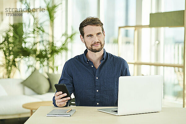 Geschäftsmann  der ein Mobiltelefon hält  während er mit einem Laptop im Büro zu Hause sitzt