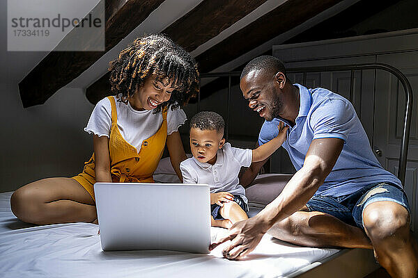 Glückliche Familie mit Sohn auf dem Bett sitzend und Laptop benutzend auf dem Dachboden zu Hause