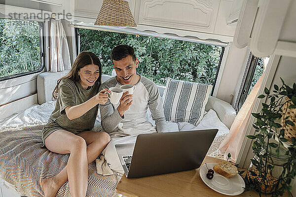Glückliches junges Paar hält Kaffeetassen und führt einen Videoanruf über einen Laptop im Wohnmobil