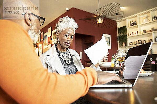 Ältere Frau diskutiert mit einem männlichen Kollegen  der im Restaurant am Laptop arbeitet