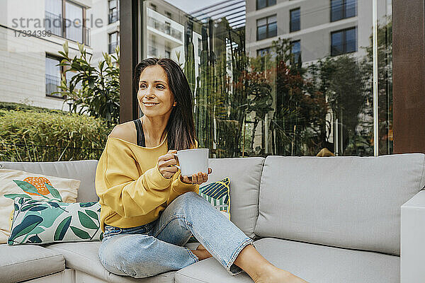 Lächelnde Frau mit Tasse  die auf dem Sofa im Hinterhof wegschaut
