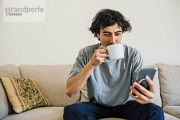 Mann trinkt Kaffee  während er zu Hause sein Mobiltelefon benutzt