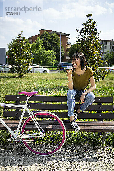 Junge Frau mit Fahrrad  die eine Sprachnachricht über ihr Smartphone sendet  während sie auf einer Bank im Park sitzt