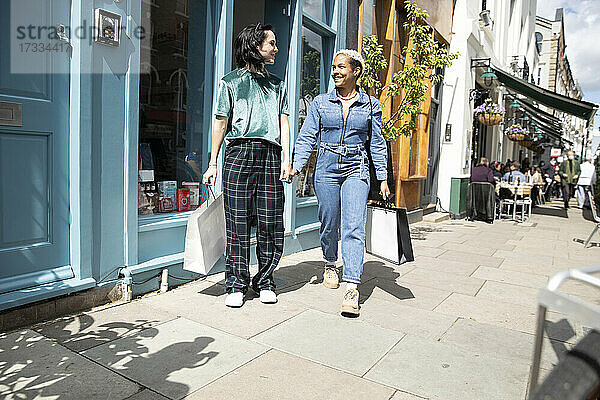 Lächelnde Frauen mit Einkaufstasche  die sich beim Gehen auf dem Fußweg an den Händen halten
