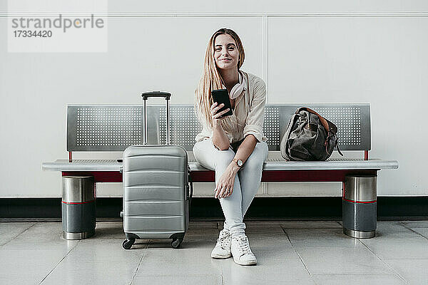 Schöne Frau  die ihr Smartphone in der Hand hält  während sie mit Gepäck auf Rädern am Bahnhof sitzt