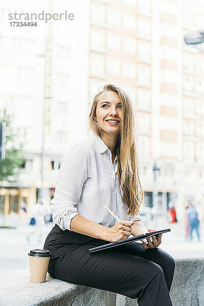 Lächelnde Geschäftsfrau mit digitalem Tablet und Einweg-Kaffeebecher auf einer Bank sitzend