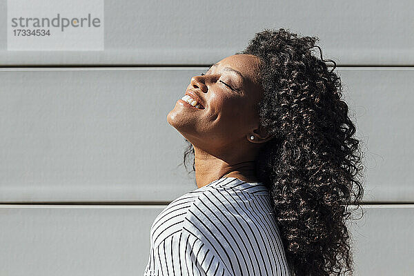 Lächelnde Frau mit geschlossenen Augen an der Wand stehend an einem sonnigen Tag