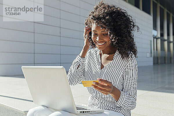 Lächelnde Frau mit Kreditkarte beim Online-Shopping  während sie vor einem Bürogebäude sitzt