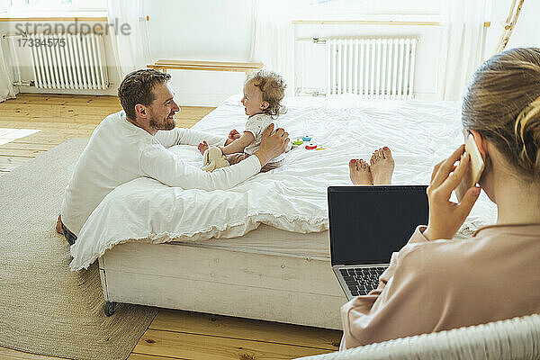 Lächelnder Vater und Tochter schauen sich an  während die Frau im Schlafzimmer mit dem Handy telefoniert