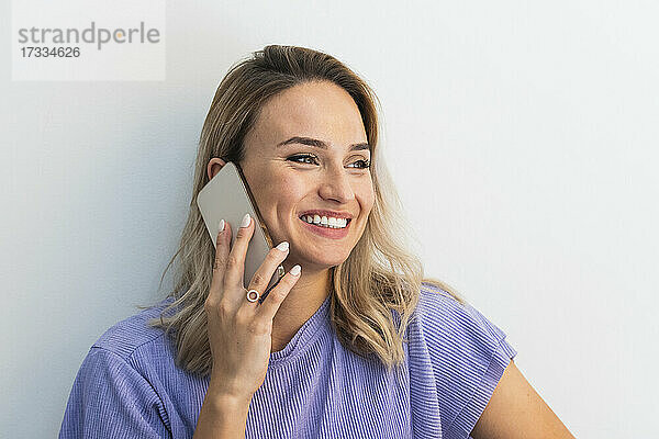 Lächelnde junge Frau  die wegschaut  während sie mit einem Handy vor einer Wand spricht