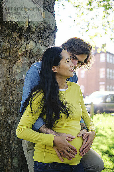 Mann umarmt schwangere Frau von hinten vor einem Baumstamm