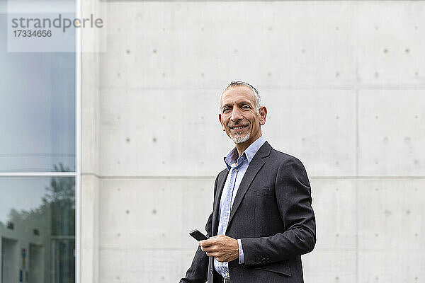 Lächelnder Geschäftsmann mit Smartphone in der Nähe eines Bürogebäudes