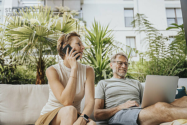 Frau telefoniert mit einem Mann  der auf der Terrasse einen Laptop benutzt