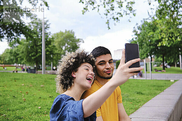 Glückliche junge Frau  die ein Selfie mit ihrem Freund über ihr Smartphone macht  während sie im Park sitzt