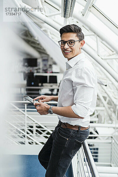 Lächelnder Geschäftsmann hält ein digitales Tablet und lehnt sich an ein Geländer