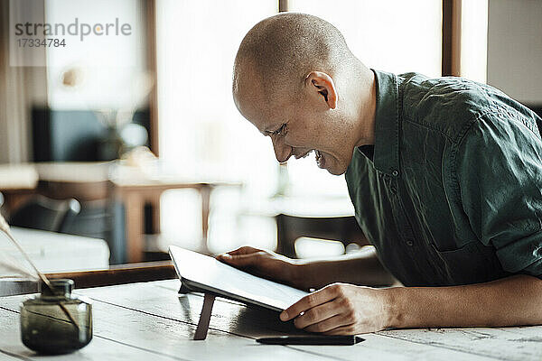 Glücklicher glatzköpfiger Geschäftsmann  der in einem Café sitzt und auf ein digitales Tablet schaut