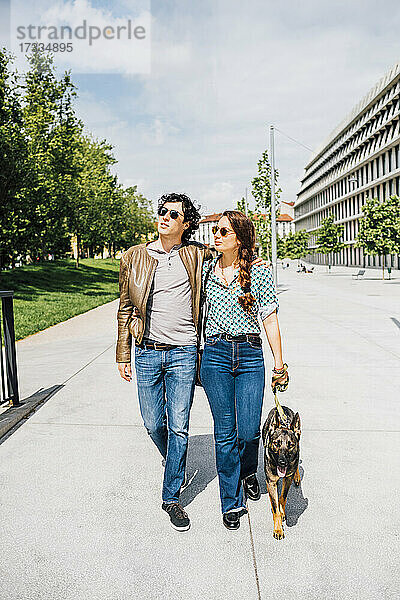 Paar  das mit seinem Hund an einem sonnigen Tag auf einer Straße in der Stadt spazieren geht