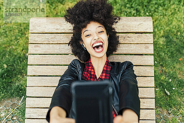 Glückliche Frau  die ein Selfie mit ihrem Smartphone macht  während sie auf einem Stuhl im Park liegt