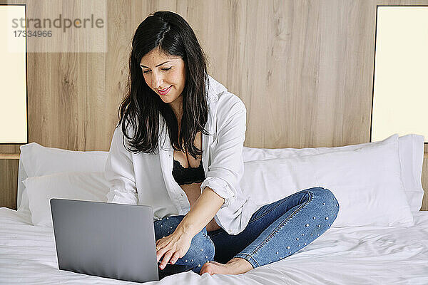 Schwangere Frau im mittleren Erwachsenenalter  die einen Laptop benutzt  während sie auf dem Bett im Schlafzimmer sitzt