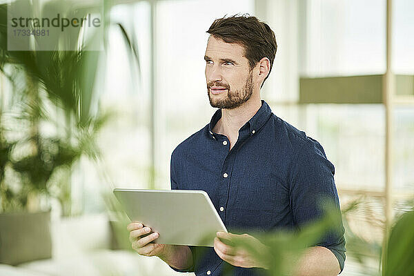 Nachdenklicher Geschäftsmann schaut weg  während er zu Hause ein digitales Tablet hält