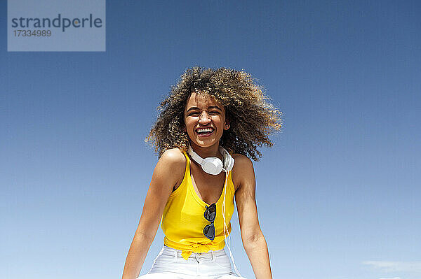 Junge Frau mit lockigem Haar lacht  während sie unter blauem Himmel an einem sonnigen Tag sitzt