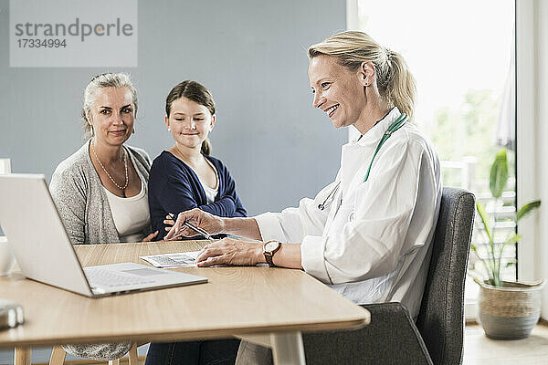 Lächelnder Kinderarzt  der auf einen Laptop schaut  während er mit einem Mädchen und einer Frau im Büro sitzt