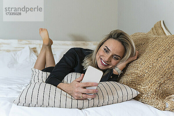 Lächelnde weibliche Fachkraft  die ein Smartphone benutzt  während sie zu Hause auf dem Bett liegt