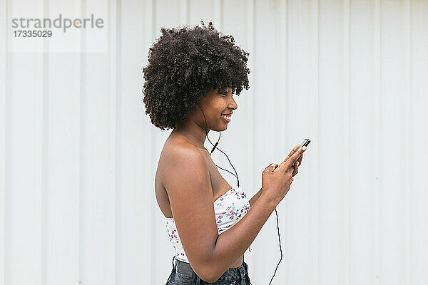 Lächelnde Afro-Frau mit Smartphone hört Musik über Kopfhörer an einer weißen Wand