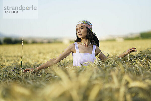 Junge schöne Frau mit Kopftuch steht in einem Weizenfeld und schaut weg