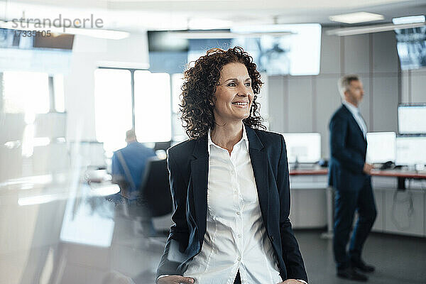 Lächelnde Geschäftsfrau  die wegschaut  während ein Kollege im Hintergrund im Kontrollraum sitzt