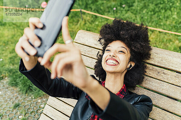Glückliche Frau  die ein Selfie mit ihrem Mobiltelefon macht  während sie auf einem Stuhl im Park liegt