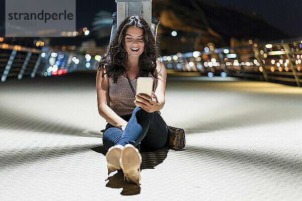 Lächelnde Frau  die ein Mobiltelefon benutzt  während sie nachts auf dem Pier sitzt