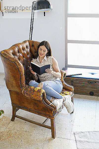 Frau liest ein Buch  während sie auf einem braunen Ledersessel im Studio sitzt