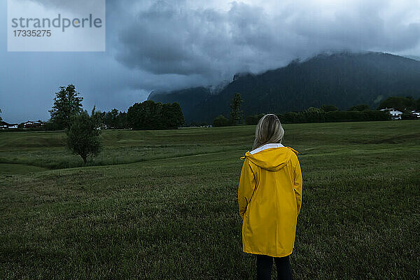 Mittlere erwachsene Frau in gelbem Regenmantel steht auf Gras unter dramatischem Himmel