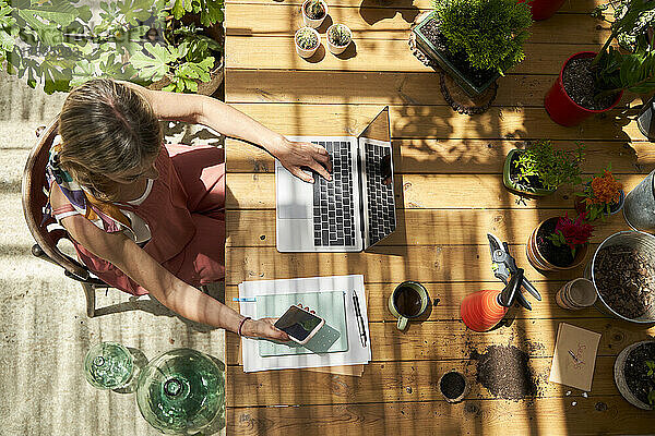 Frau  die ein Smartphone benutzt  während sie mit einem Laptop an einem Tisch im Hinterhof sitzt