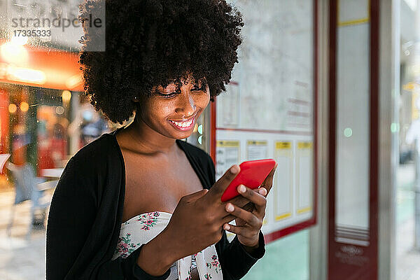 Lächelnde Afro-Frau  die an einer Bushaltestelle eine SMS über ihr Mobiltelefon schreibt