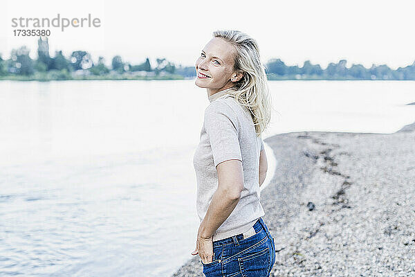 Lächelnde Frau mit Hand in der Tasche am Flussufer stehend