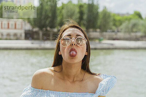 Frau mit Sonnenbrille streckt vor einem Fluss die Zunge heraus