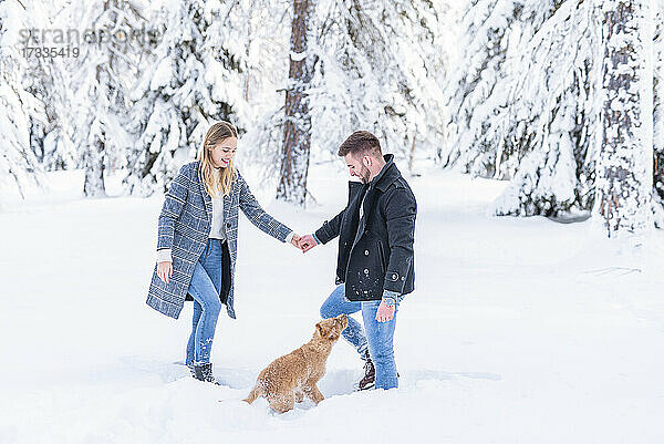 Paar hält sich an den Händen und betrachtet den Hund im Schnee während des Urlaubs