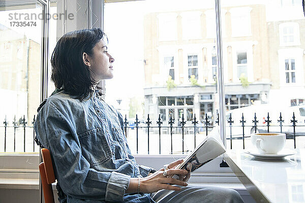 Junge Frau sitzt mit einem Buch im Café und schaut durch das Fenster