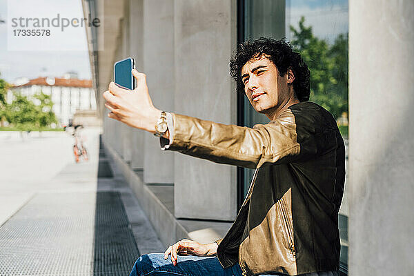 Junger Mann  der ein Selfie mit seinem Smartphone macht  während er im Freien sitzt
