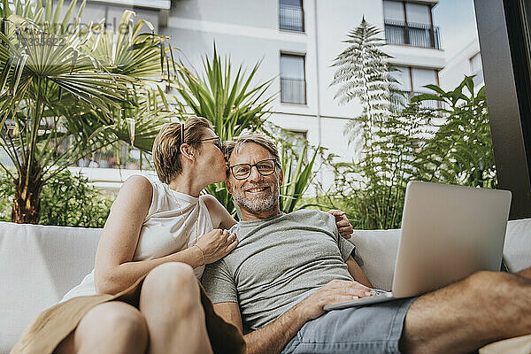 Frau küsst Mann  der mit Laptop auf einem Sofa auf der Terrasse sitzt