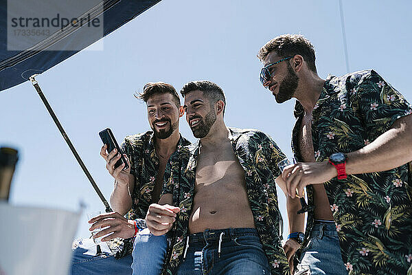 Lächelnder Mann teilt Handy mit männlichen Freunden auf einer Yacht an einem sonnigen Tag im Urlaub