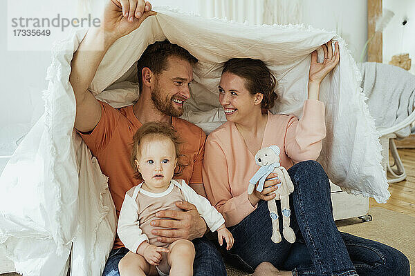 Lächelnder Mann und lächelnde Frau sitzen mit ihrer in eine Decke gehüllten Tochter zu Hause