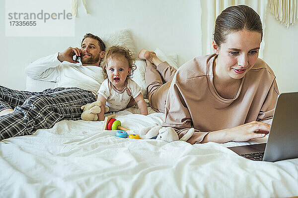 Frau benutzt Laptop  während der Mann mit seiner Tochter auf dem Bett zu Hause telefoniert