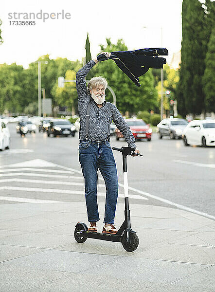 Glücklicher reifer Mann schwingt Jacke beim Rollerfahren auf der Straße