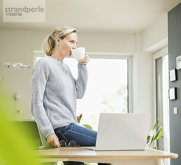 Freiberuflerin trinkt Kaffee und sitzt am Laptop auf dem Schreibtisch